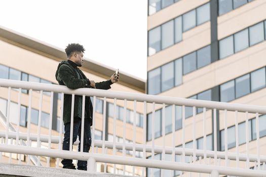 serious african black man with earphones making video call with a mobile phone leaning on a handrail outdoors in the city, technology and lifestyle concept using internet electronic device