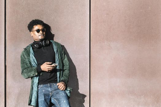 cool african black guy with casual modern clothes and a mobile phone and headphones to listen music leaning against wall, technology and lifestyle concept