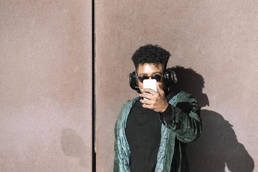 cool african black man with casual modern clothes and headphones to listen music taking a selfie with the phone leaning against wall, technology and lifestyle concept