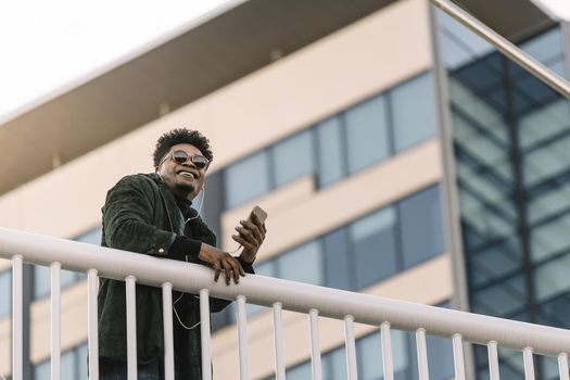 portrait of a smiling young black man with sunglasses listening to music with earphones and mobile phone while leaning on a handrail outdoors in the city, technology and lifestyle concept