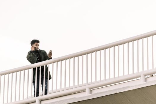 african black man with headphones making video call with a mobile phone leaning on a handrail outdoors in the city, technology and lifestyle concept using internet electronic device