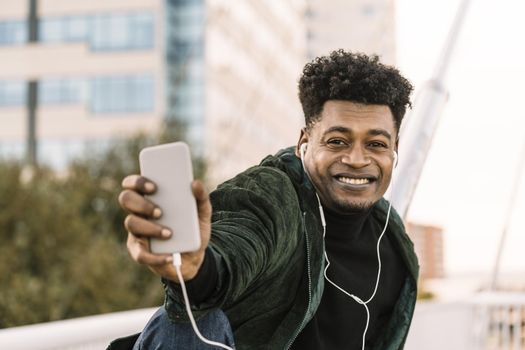 handsome smiling young african black man making a selfie or a video call with smartphone and earphones while sitting in the city, lifestyle and technology concept using internet electronic device