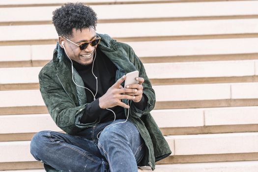 black young male in casual modern clothes with phone smiling and sitting on stairs while making a video call with earphones, technology and lifestyle concept