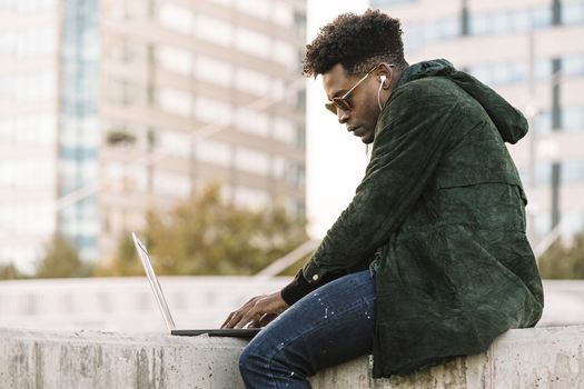 handsome young student with sunglasses and earphones working on laptop sitting outdoors in the city, lifestyle and technology concept using internet electronic device