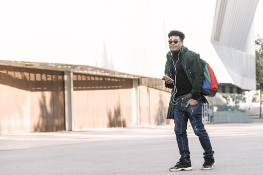 black young man with earphones walking by the city and listening music with a mobile phone, technology and lifestyle concept using internet electronic device outdoors