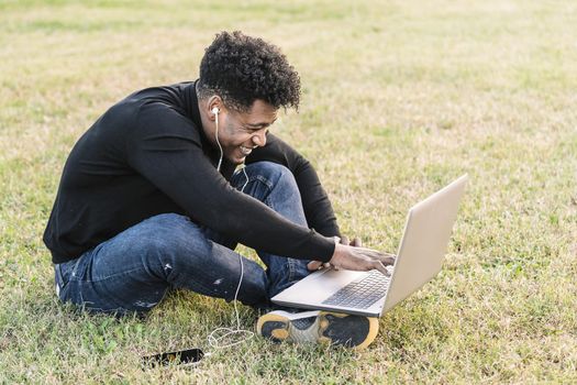 smiling young black man laughing with white earphones and phone working typing in a computer sitting on the lawn at park, lifestyle and technology concept using internet electronic device
