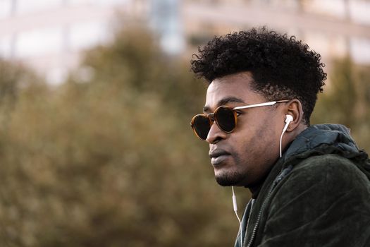 handsome young black african man with green jacket and sunglasses listening music with white earphones, city lifestyle concept