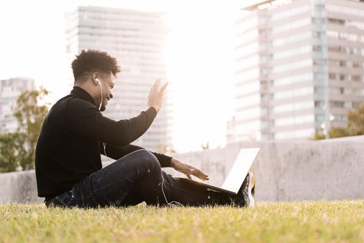 smiling young black man laughing with white earphones making a video call with the laptop sitting on the lawn in the city at sunset, lifestyle and technology concept using internet electronic device