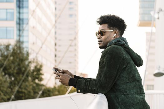cool young african black man with sunglasses listening music on smartphone with earphones while leaning on a handrail outdoors in the city, lifestyle and technology concept, copy space for text