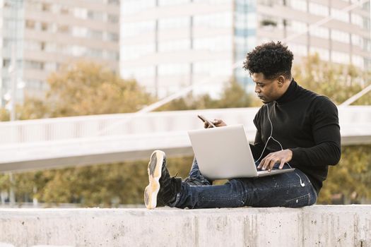 handsome black student with white earphones working with computer and smart phone sitting outdoors in the city at sunset, lifestyle and technology concept using internet electronic device