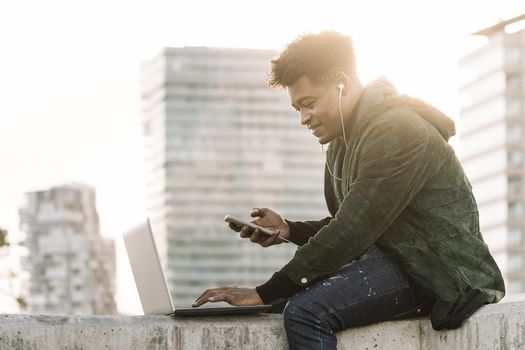 handsome young student with white earphones and phone working typing in a computer sitting outdoors in the city at sunset, lifestyle and technology concept using internet electronic device