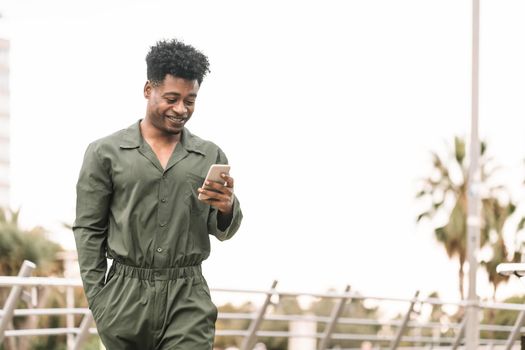 handsome young african black man smiling and texting message on smartphone while walks in a park outdoors in the city, lifestyle and technology concept, copy space for text