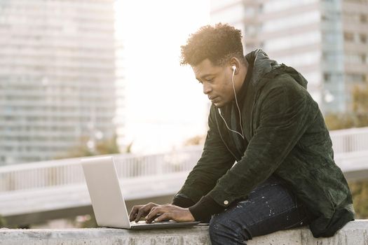 handsome young student with white earphones working typing in a laptop sitting outdoors in the city at sunset, lifestyle and technology concept using internet electronic device