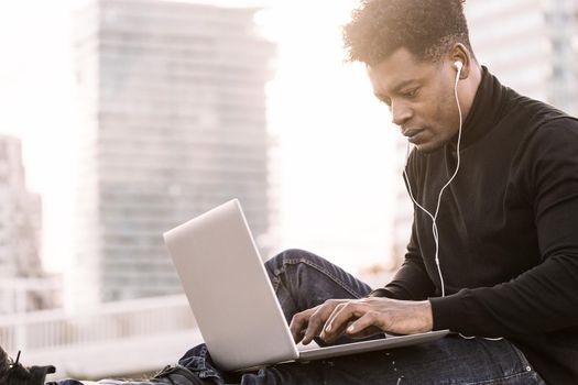 handsome black student with white earphones working typing in a computer sitting outdoors in the city at sunset, lifestyle and technology concept using internet electronic device