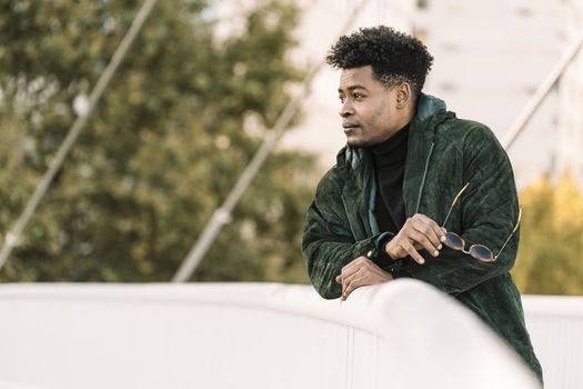 attractive young african black man with green jacket and sunglasses while leaning on a handrail outdoors in the city, lifestyle concept