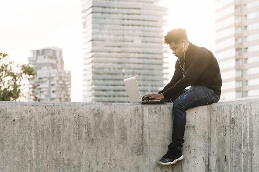 handsome black man with white earphones working typing in a computer sitting outdoors in the city at sunset, lifestyle and technology concept using internet electronic device
