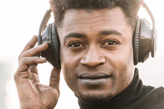 portrait of a happy african black man smiling listening music with a hand touching the headphones at sunset, technology and lifestyle concept