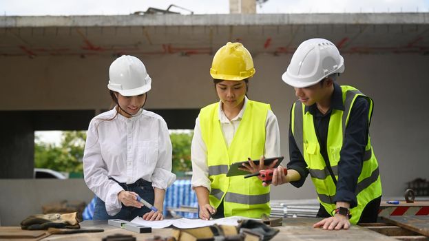 Architect team in safety helmet using digital tablet and working with blueprints during inspecting construction site.