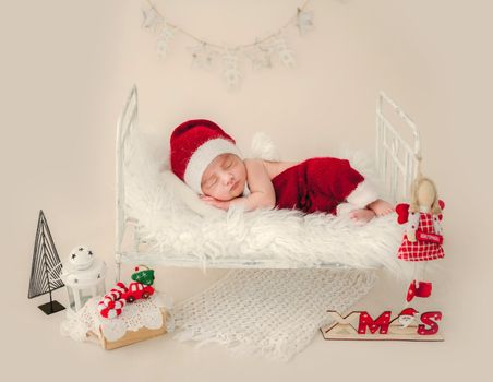 Newborn baby boy wearing Santa costume and hat sleeping in tiny bed on white fur with Christmas decoration. Infant child kid New Year holiday napping portrait