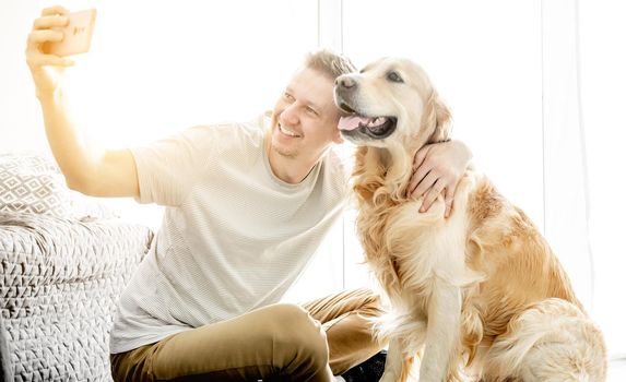 A young handsome man with his golden retriever dog takes a selfie in a bright living room.