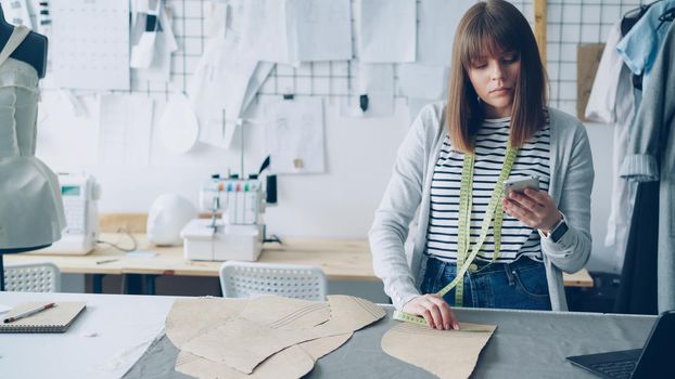 Young seamstress is checking clothing paper patterns and measuring them with tape-measure while looking at smart phone. Nice light studio with garments, textiles and sewing machine in background.