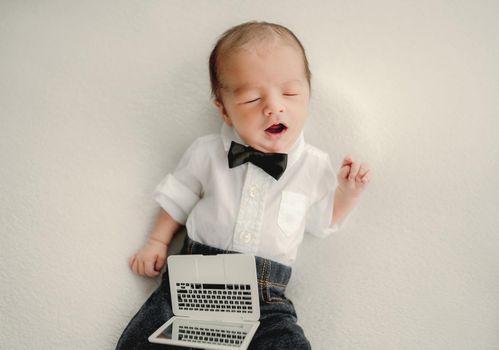 Newborn baby boy wearing festive costume with bow-tie and holding toy laptop falling asleep and yawning. Infant child kid office studio portrait