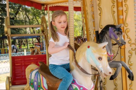 Close-up portrait of a blonde girl of European appearance in white T-shirt. A child rides colorful carousel with horses in an amusement park. Summer day. The concept of happiness, family day, holiday