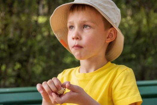 Close-up portrait of a European-looking boy eating popcorn in the park. The concept of a hungry child, malnutrition, snacking, unhealthy food. Summer day, a boy in a light panama hat and a yellow T-shirt.