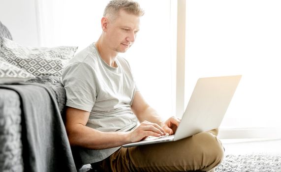 A young handsome man works at a laptop in a bright living room.