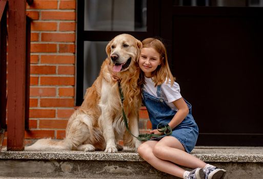 Preteen girl wearing jeans dress with golden retriever dog sitting outdoors in summertime. Pretty kid hugging purebred fluffy doggy pet in city