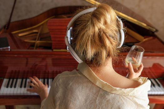 A young blonde girl, in white headphones, sits at the piano with her back. A woman holds a glass of white wine in her hands. The concept of loneliness, depression, alcoholism. Life style, close-up
