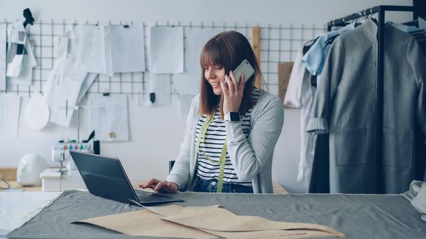 Small business owner is ordering fabric for her tailor's shop with mobile phone and working with laptop. Woman is talking and typing simultaneously. Modern means of communication concept.