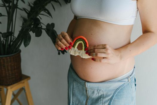 A pregnant girl in a denim jumpsuit holds a rainbow in her hands for the decor of a children's room. Great time to have a baby. Future mom. Rainbow macrame.