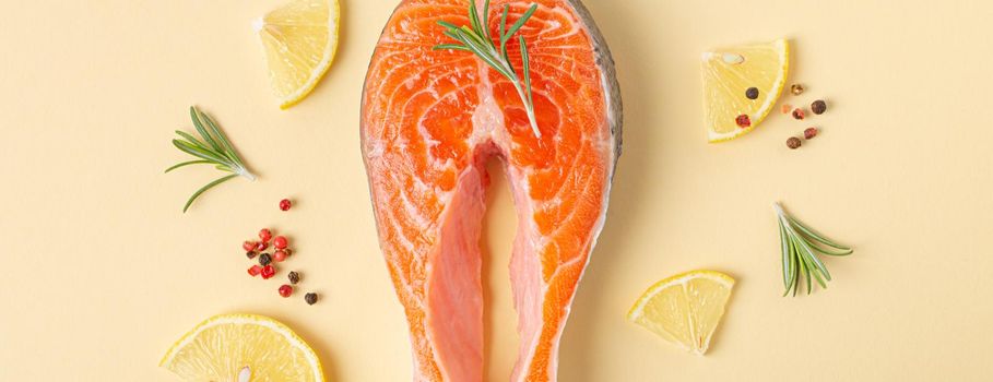 Uncooked raw fresh fish salmon steak top view on beige pastel background with rosemary, lemon wedges and spices, delicacy healthy fish cooking and nutrition concept flat lay