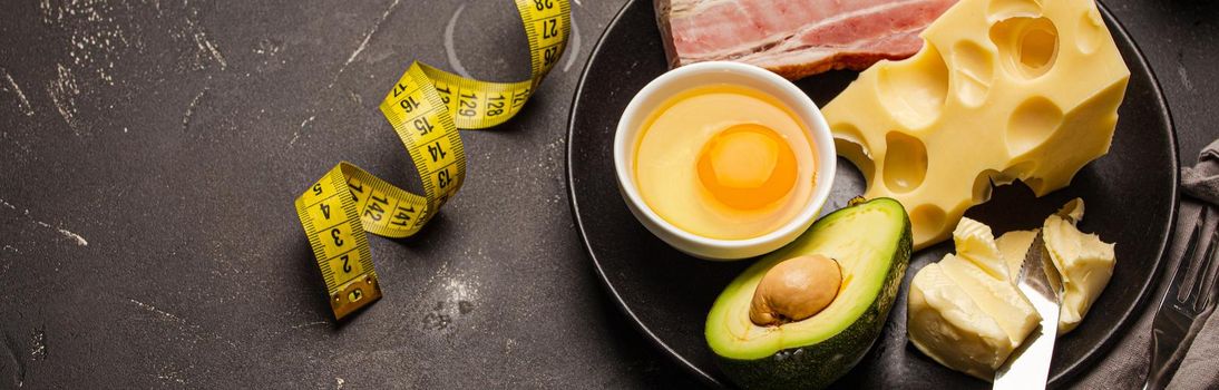 Keto foods as butter, olive oil, fried egg, avocado, fat meat bacon, cheese for ketogenic diet on black ceramic plate, measure tape on dark rustic stone background, products for weight loss copy space