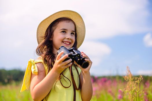 Happy cute girl 4-5 years old, in a straw hat and a yellow t-shirt, stands in a field with high grass against a blue sky in the summer, holds binoculars. Copy space