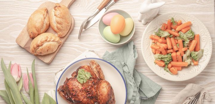 Easter food on white rustic table: pastel colored eggs, roasted chicken and vegetables, buns and spring flowers tulips top view flay lay, Easter family dinner meal with festive dishes