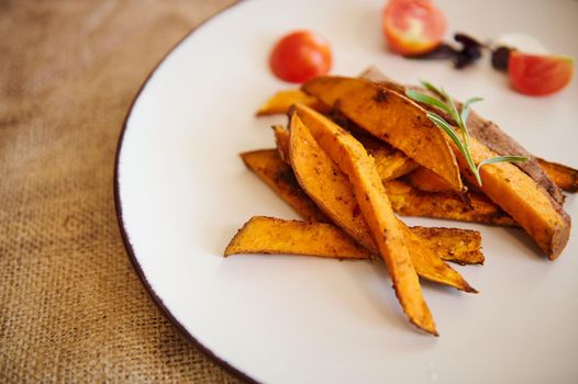 Close-up of a white ceramic plate with wedges of roasted organic sweet potato with rosemary in a table with linen tablecloth. Healthy vegetarian food. Vegan's Day. Restaurant menu