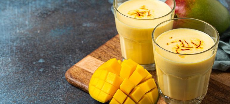 Healthy Indian Ayurveda drink mango lassi in two glasses on rustic concrete table with fresh ripe cut mango, yellow blended beverage made of mango fruit, yoghurt or milk curd and spices copy space