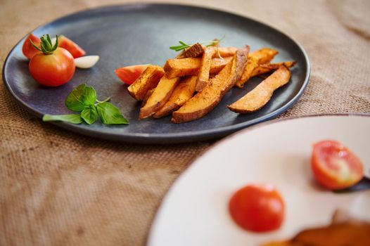 Homemade cooked sweet potatoes with spices and herbs, served with rosemary and cherry tomatoes on blue ceramic plate. Close-up. Roasted batata. Delicious healthy vegan food