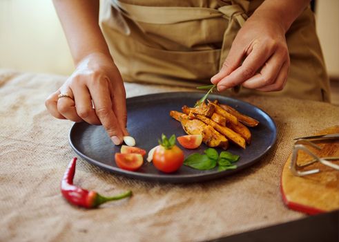 Close-up of the hands of a housewife or chef decorating dishes of fried sweet potato with cherry tomatoes and herbs, preparing delicious healthy vegan meal for lunch.