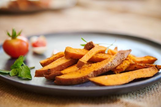 Homemade cooked sweet potatoes with spices and herbs, served with rosemary and cherry tomatoes on blue ceramic plate. Close-up