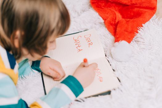 Close up hands of Kid boy, lying on couch and writing letter in note pad to dear Santa at home in Christmas holidays. Child wish list. Dreams of a Christmas gifts. Merry Christmas and Happy new year