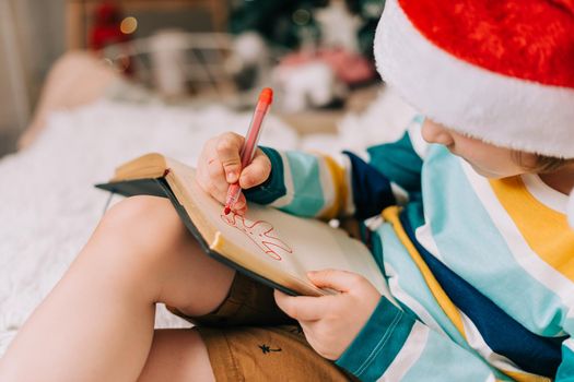 Kid boy in red Santa hat writing the letter and drawing to dear Santa at home. Child wish list. Dreams of a Christmas gifts. Merry Christmas and Happy new year