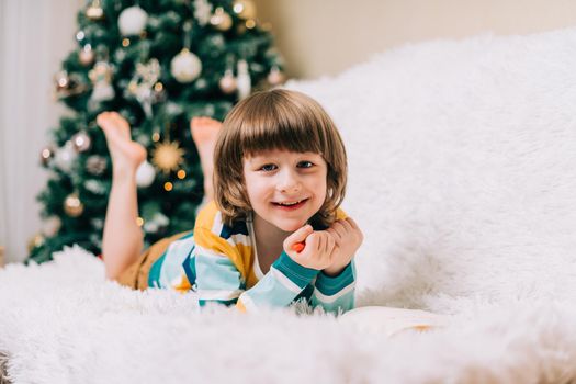 Smiling Kid boy lying on couch and writing the letter to dear Santa at home near Christmas tree. Child wish list. Dreams of a Christmas gifts. Merry Christmas and Happy new year