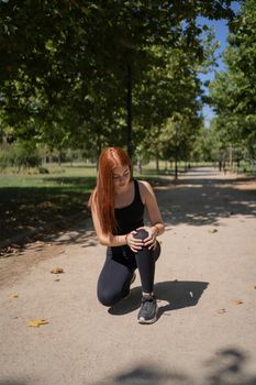 Full body redhead young sportswoman touching dislocated kneecap while kneeling on path during running session on sunny summer day in park