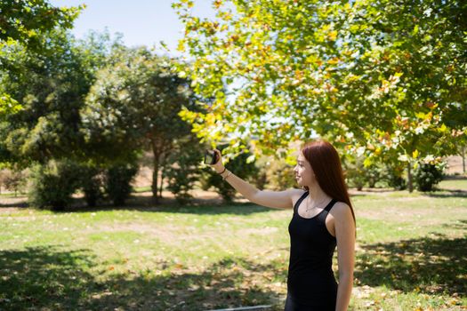 Glad young woman in casual clothes smiling and taking selfie via smartphone while standing on sunlit park alley on summer weekend day