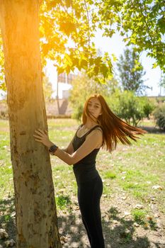 Young female athlete tossing long redhead hair and looking at camera while touching tree trunk during break in running session on sunny day in park