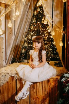 A girl in a festive light dress next to a Christmas tree, lights of garlands in the background. The concept of New Year holidays.