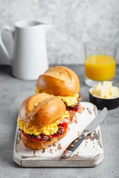 Breakfast sandwiches with scrambled egg, bacon, cheese, tomato on white wooden board, glass with fresh orange juice, white background. Making breakfast concept, selective focus